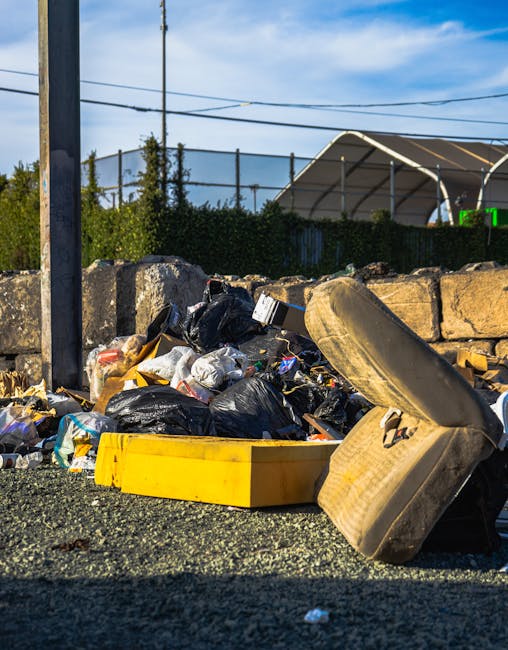 A collection of discarded waste materials is situated on a gravel surface outdoors next to a low stone wall. The rubbish includes black plastic trash bags, a yellow plastic container, an old, dirty ca