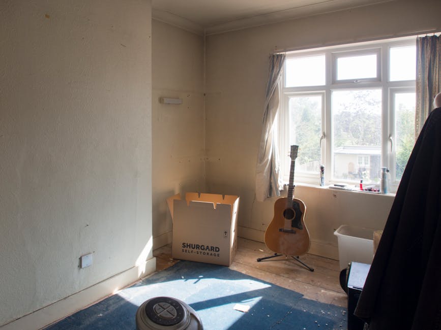 Inside a minimally furnished room with cream-colored walls and a large window allowing natural daylight to illuminate the space. Near the window, there is an acoustic guitar standing upright on a blac